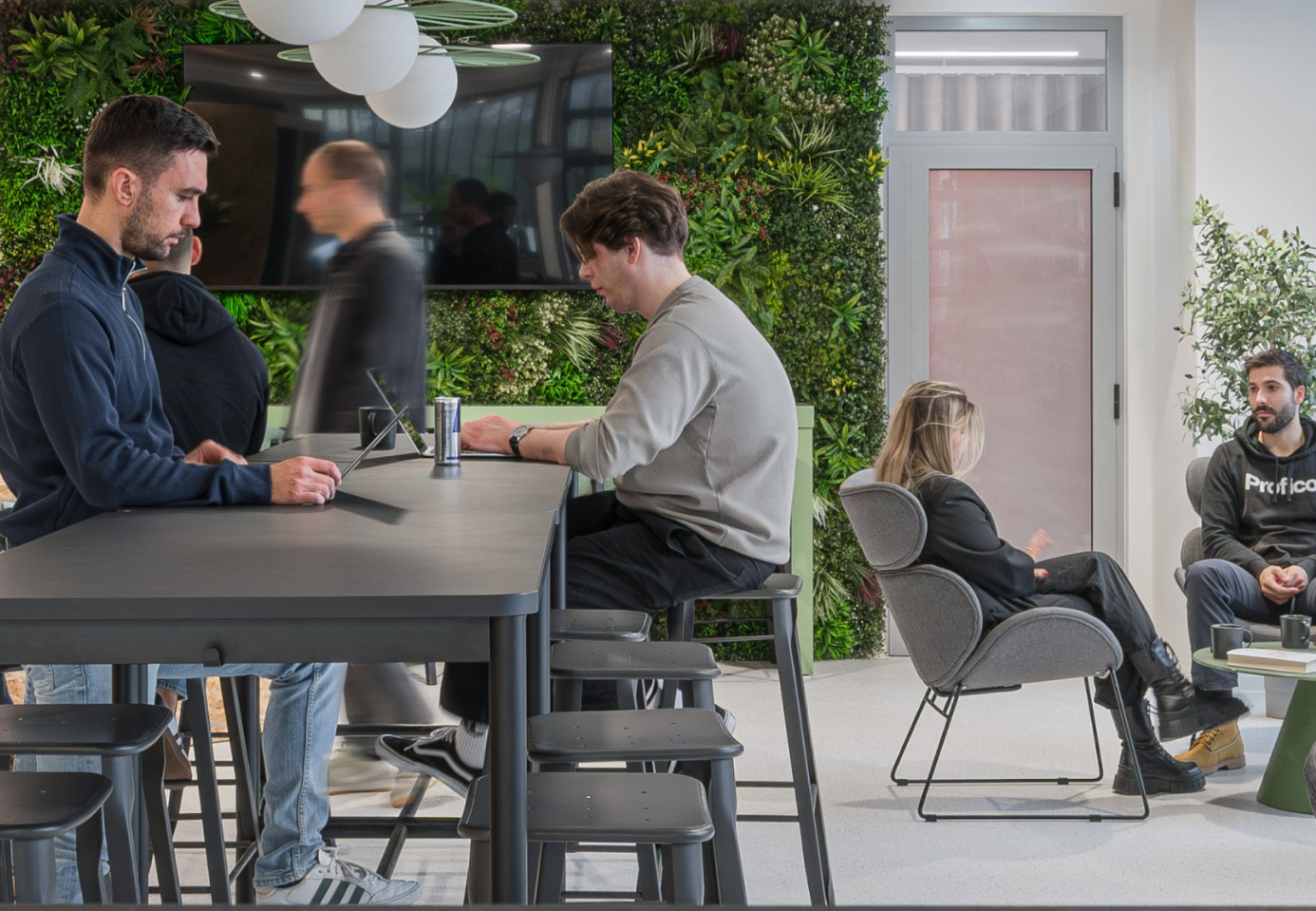 A modern, open-plan office environment where several people are working. Two men are focused on laptops at a high black communal table in the foreground, with a blurred figure walking by in the background. The office features a prominent living green wall with an embedded monitor. On the right side, a man in a black hoodie and a woman sit in lounge chairs, appearing to have a conversation.