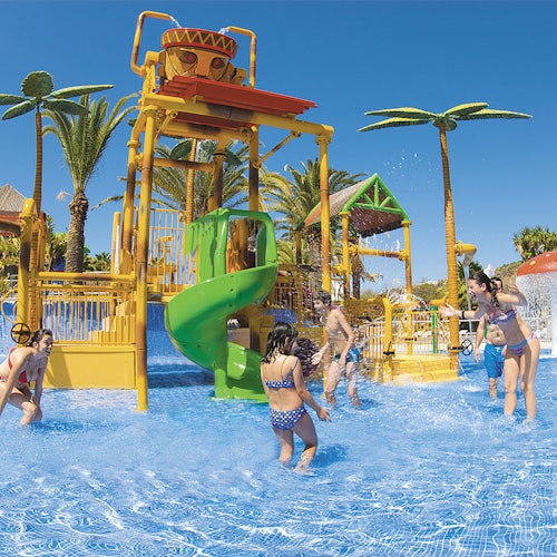Children playing in a water park with slides and splash features; palm trees surround the area under a clear blue sky.