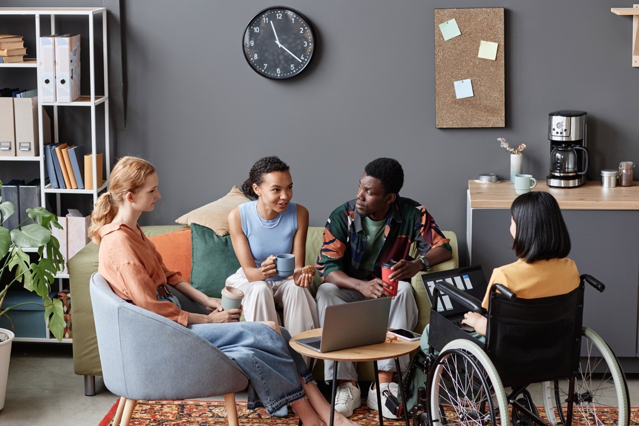 Four people, including one person in a wheelchair, sit in a casual office setting, talking and holding mugs. A laptop is on a small table, with a clock, bulletin board, and coffee maker in the background.