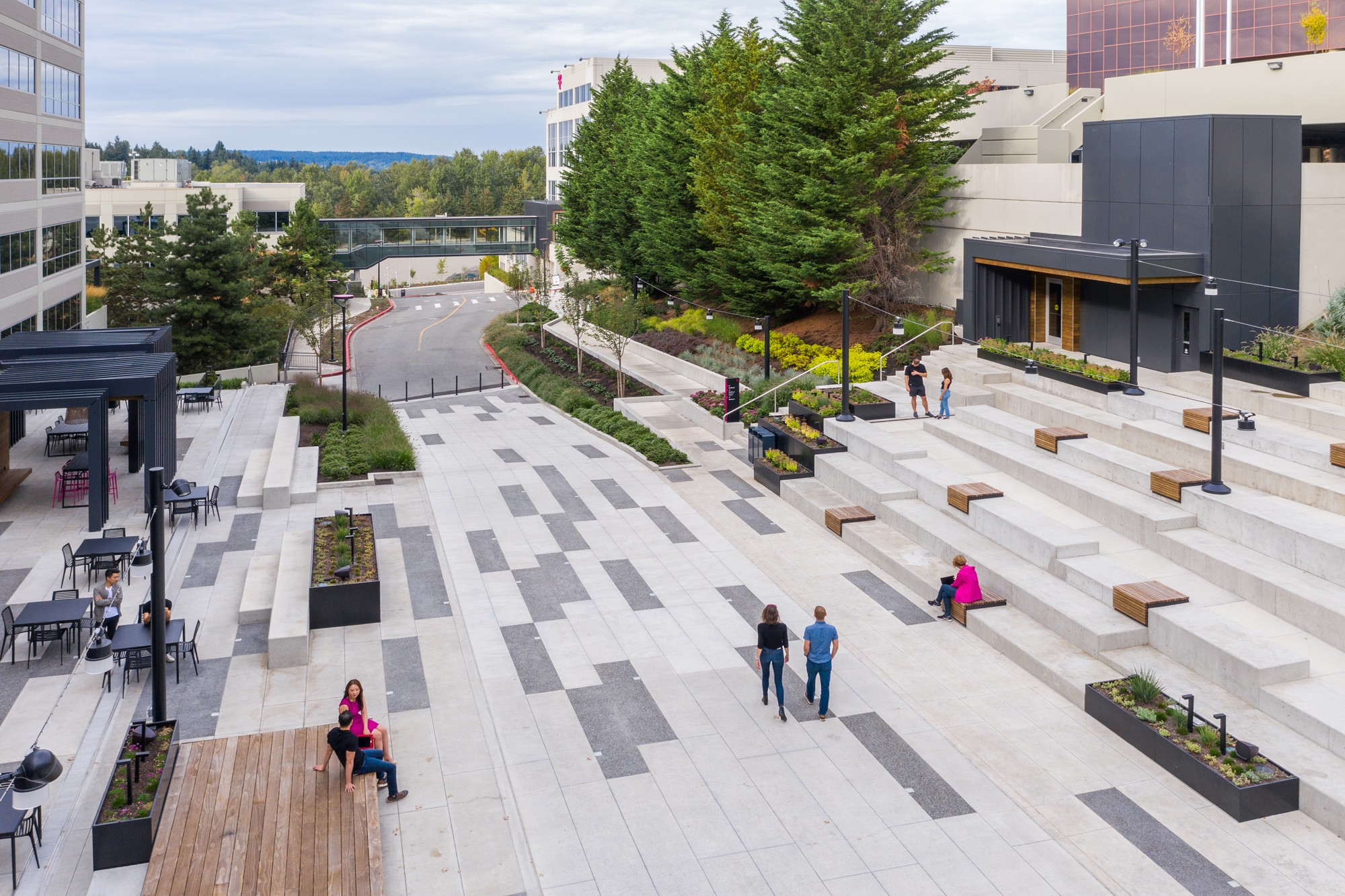 A midday aerial view of the plaza center, showcasing the wooden hybrid bench/stage, flexible seating arrangements, and a ramp gracefully weaving through the amphitheater.