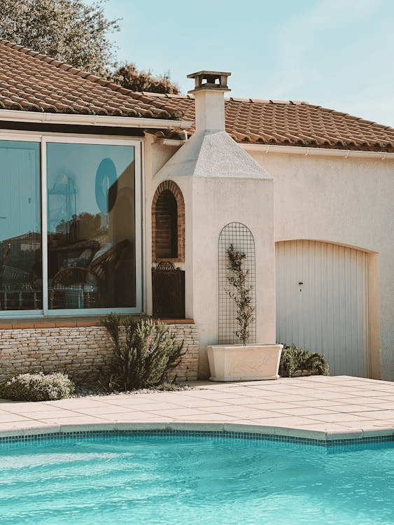 Contemporary white stucco house with large arched doorway, garage, and palm trees, featuring a pool area on a tiled patio.