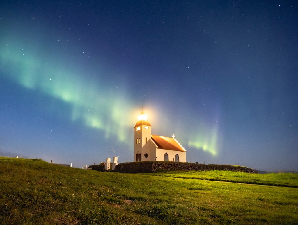 A chapel on top of a grassy hill with the northern lights glowing in the sky