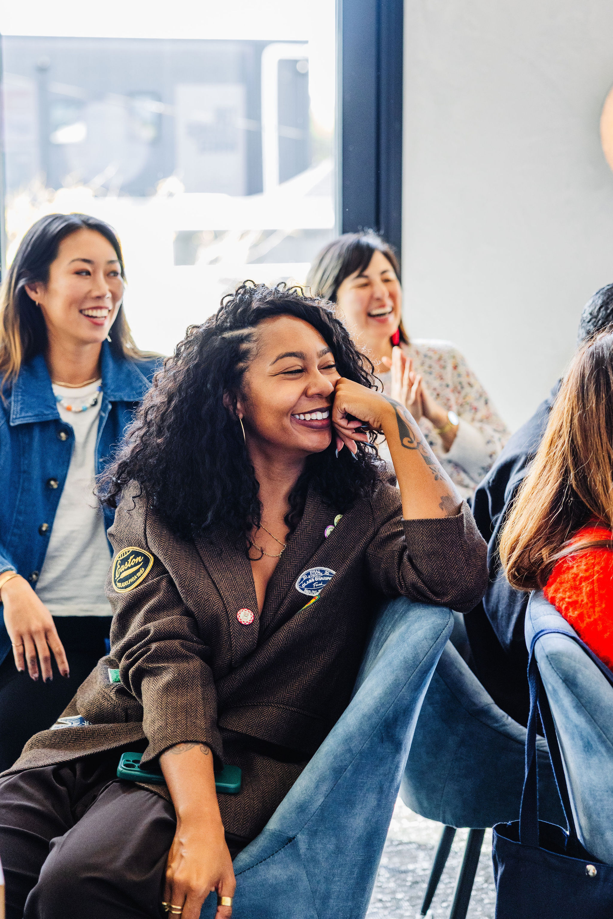 Woman with curly hair smiling and resting her chin on her hand while laughing with others in a casual group setting.