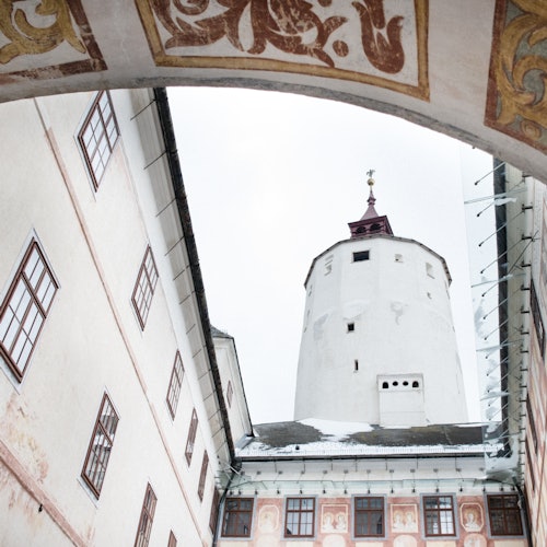 A white tower with a red roof viewed through an archway, surrounded by buildings with multiple windows and decorative details.