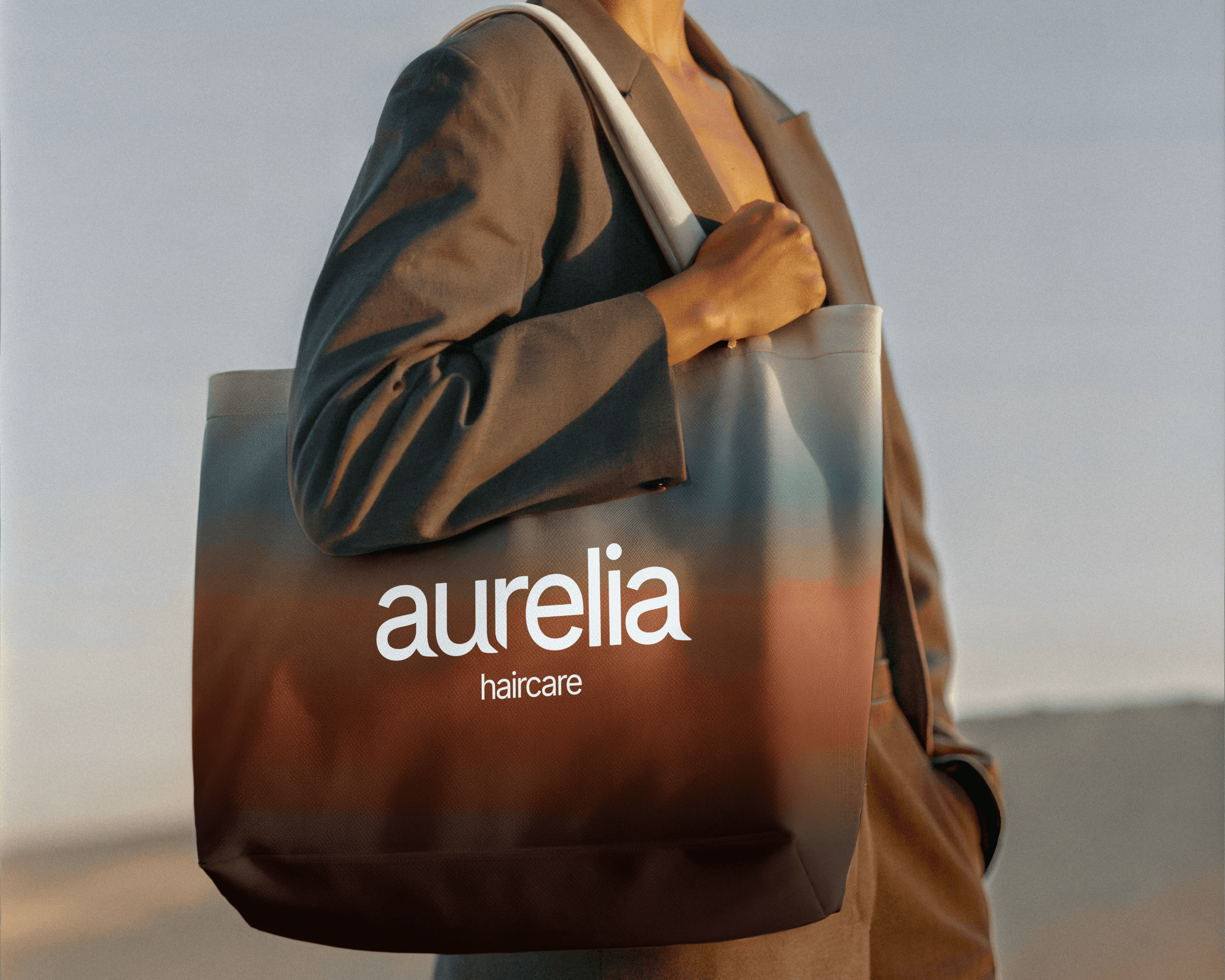 A person wearing a beige suit holding a tote bag on their shoulder. The bag has a blue and brown gradient with the logo for Aurelia Haircare on it.
