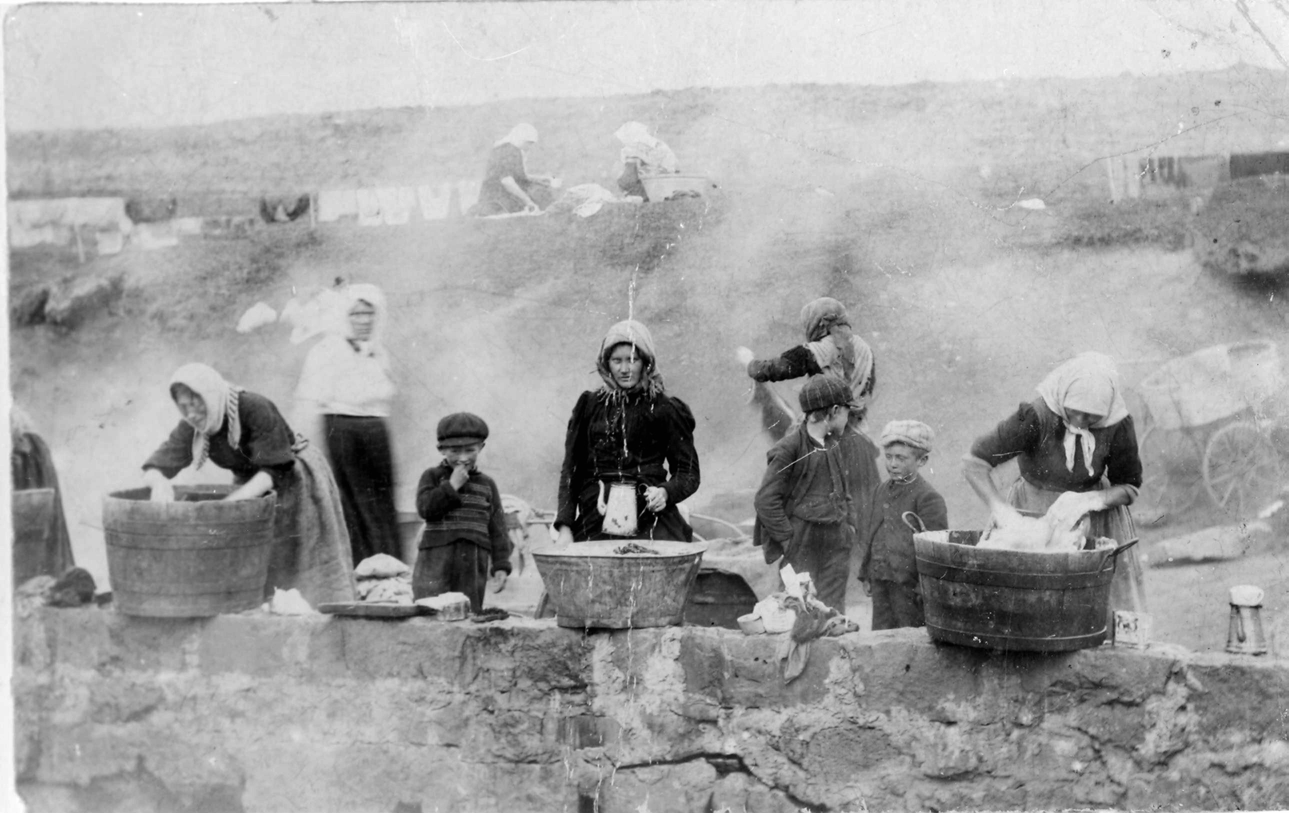Black-and-white photo of women and children doing laundry at the geothermal hot springs in Laugardalur, Reykjavík, Iceland, circa late 19th century. The women are washing clothes in large wooden tubs while steam rises from the hot spring. Children stand nearby, observing or assisting.