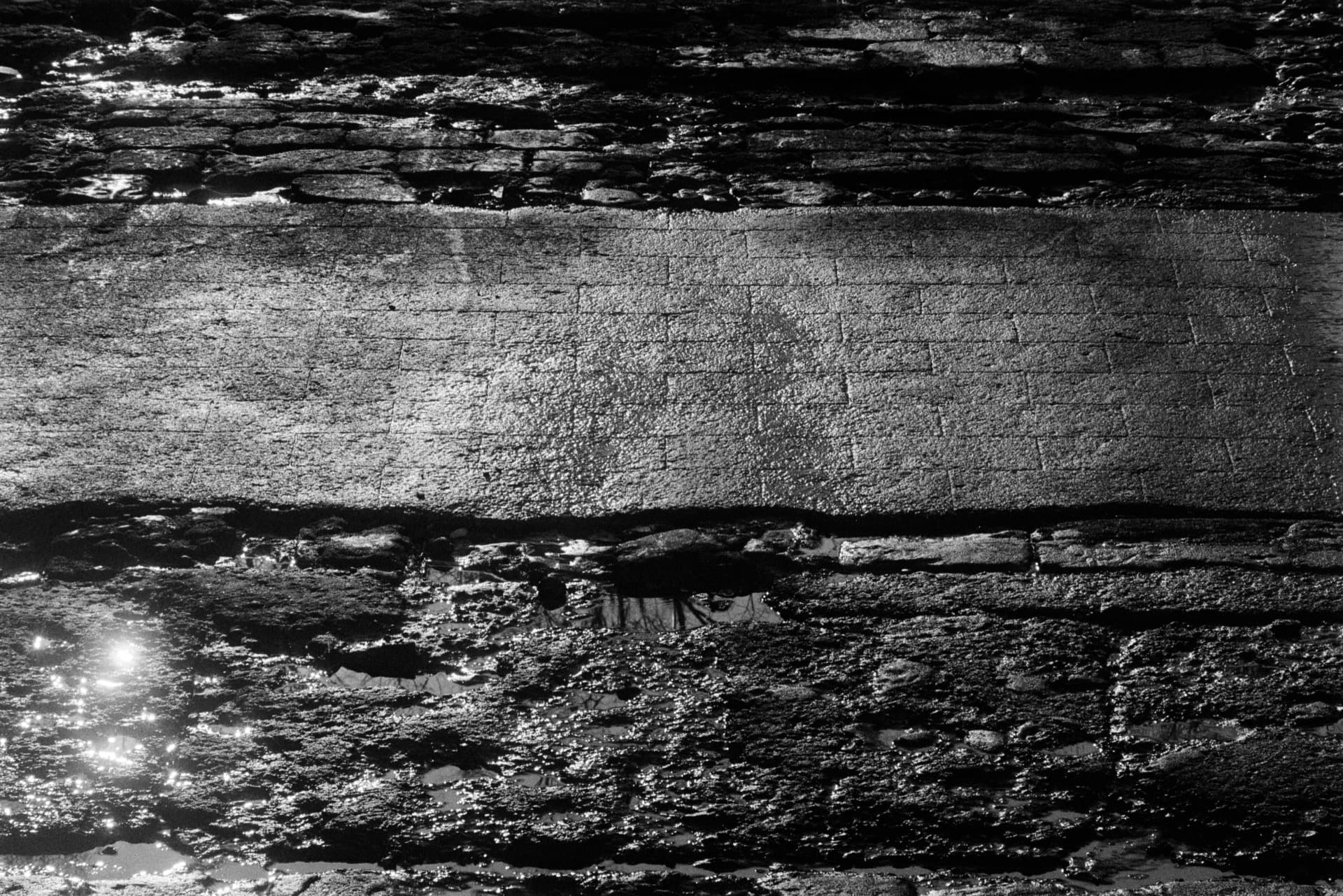 Close detail of concrete slipway leading to Thames, sunlit band across center showing weathered texture, dark water pools above and below with reflected light