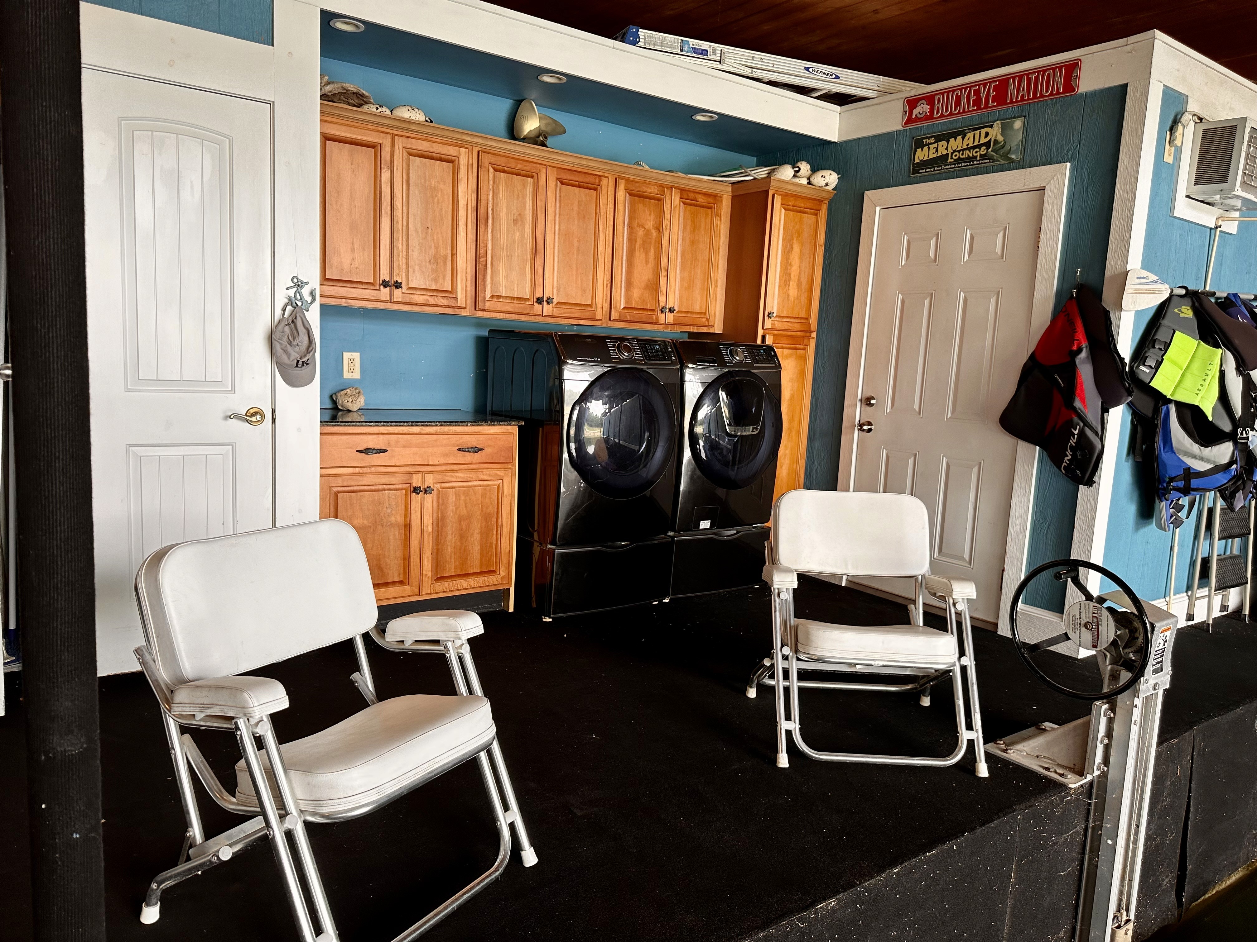 This image shows a laundry area featuring a sleek black washer and dryer set against a wall of warm wooden cabinets, accompanied by two white folding chairs on a dark carpeted floor, alongside a brightly decorated wall with life jackets and a "Mermaid" sign overhead.