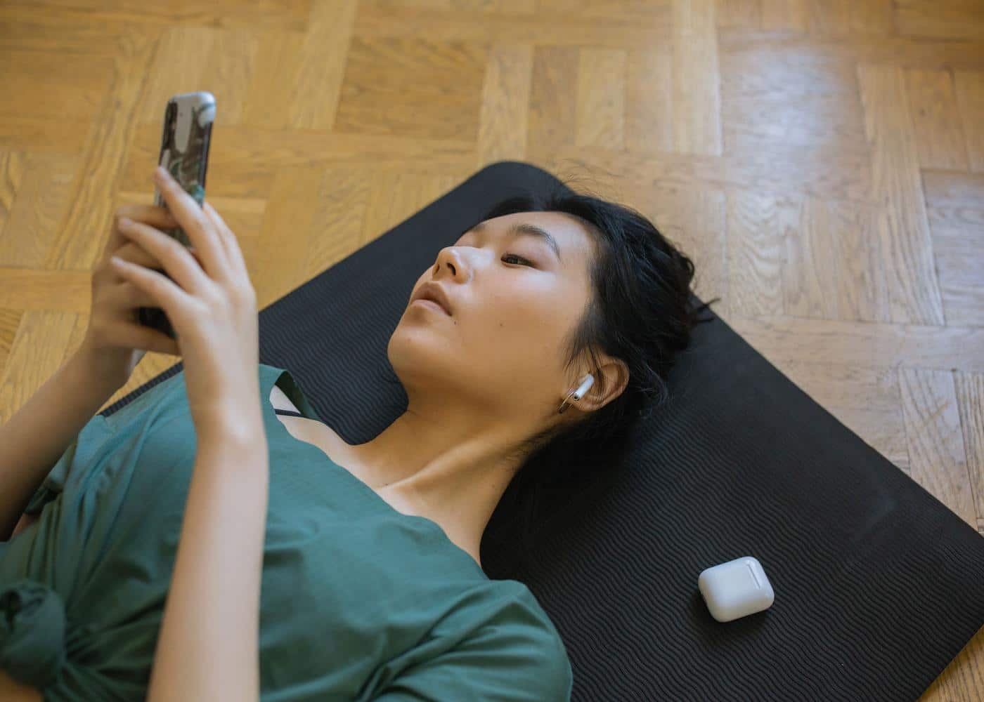 Woman laying on black yoga mat looking at smartphone with Airpods next to her
