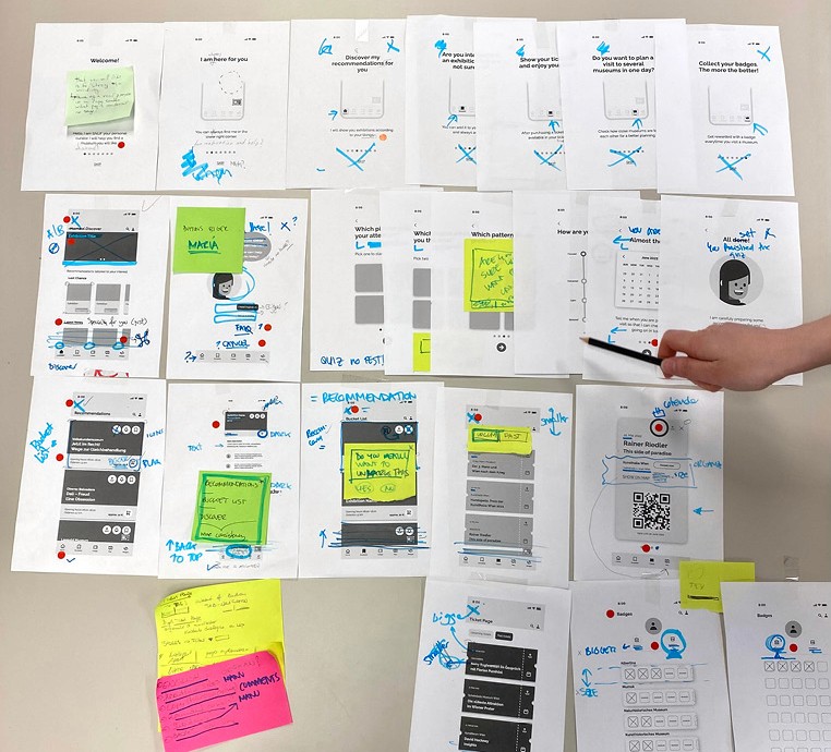 Ausgedruckte Screens mit Post Its und Korrekturen, Stift zeigt auf einen Sceeen