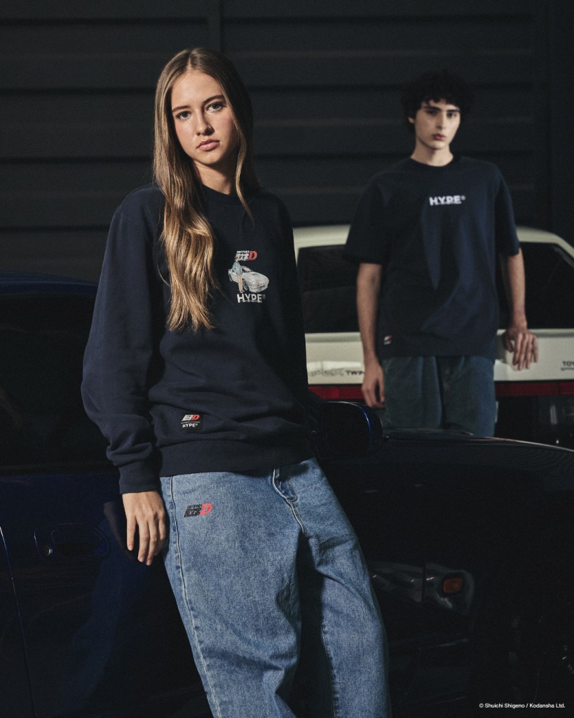 A male and female model pose in casual streetwear featuring designs inspired by Initial D, set against a backdrop of sports cars in a dimly lit environment.