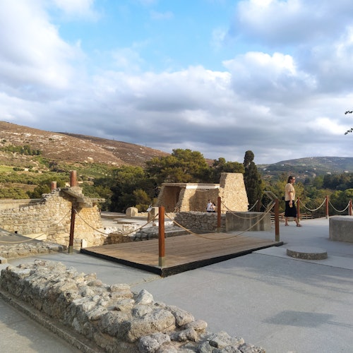 An ancient ruins site with stone structures, a wooden walkway, and two people walking against a backdrop of hills and cloudy sky.