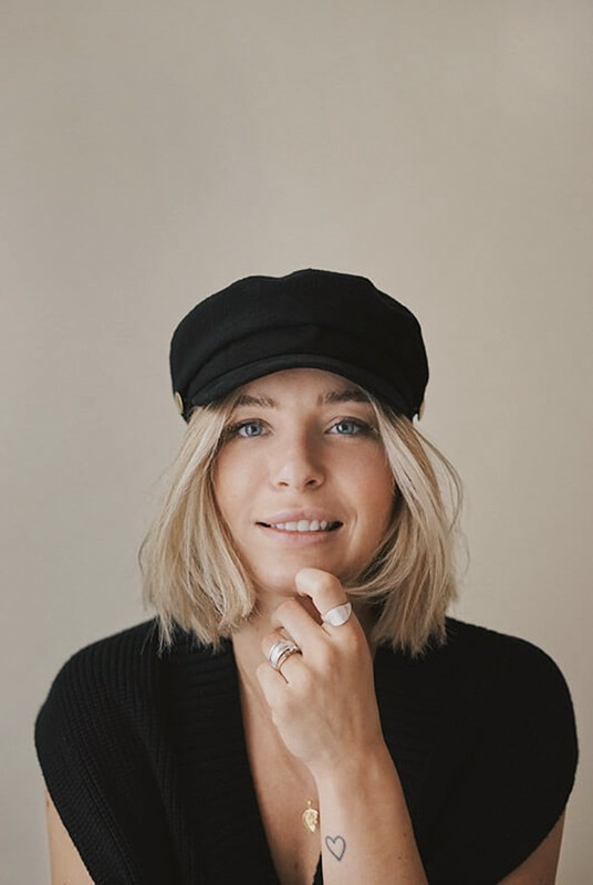 A person with shoulder-length blonde hair wearing a black newsboy cap and a black sweater.