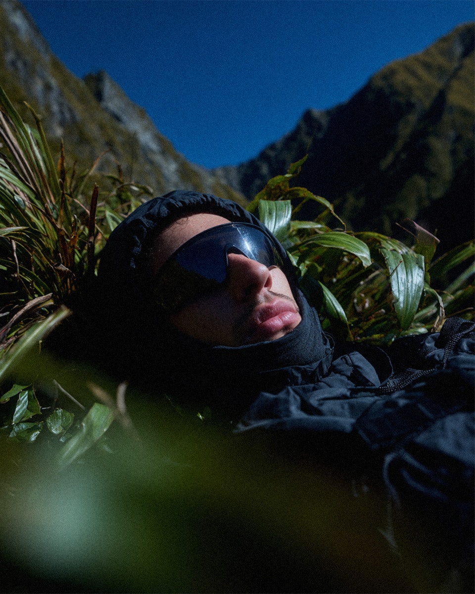 Person wearing a black hooded jacket and sunglasses lying in green grass with mountains in the background.