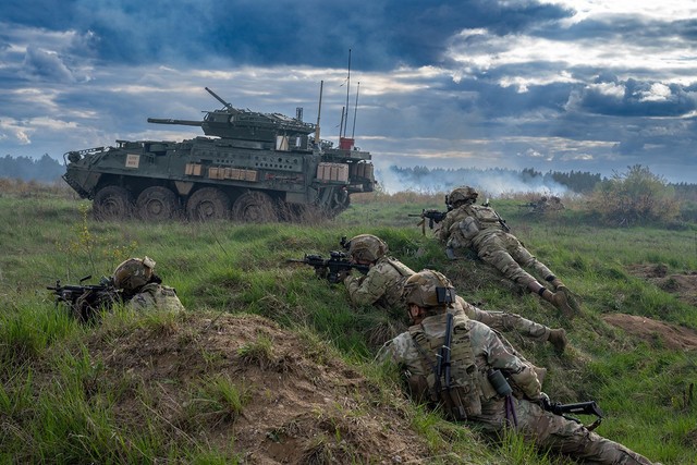 What to bring to the field — infantry soldiers prone in the grass on a field training exercise with a Stryker behind them