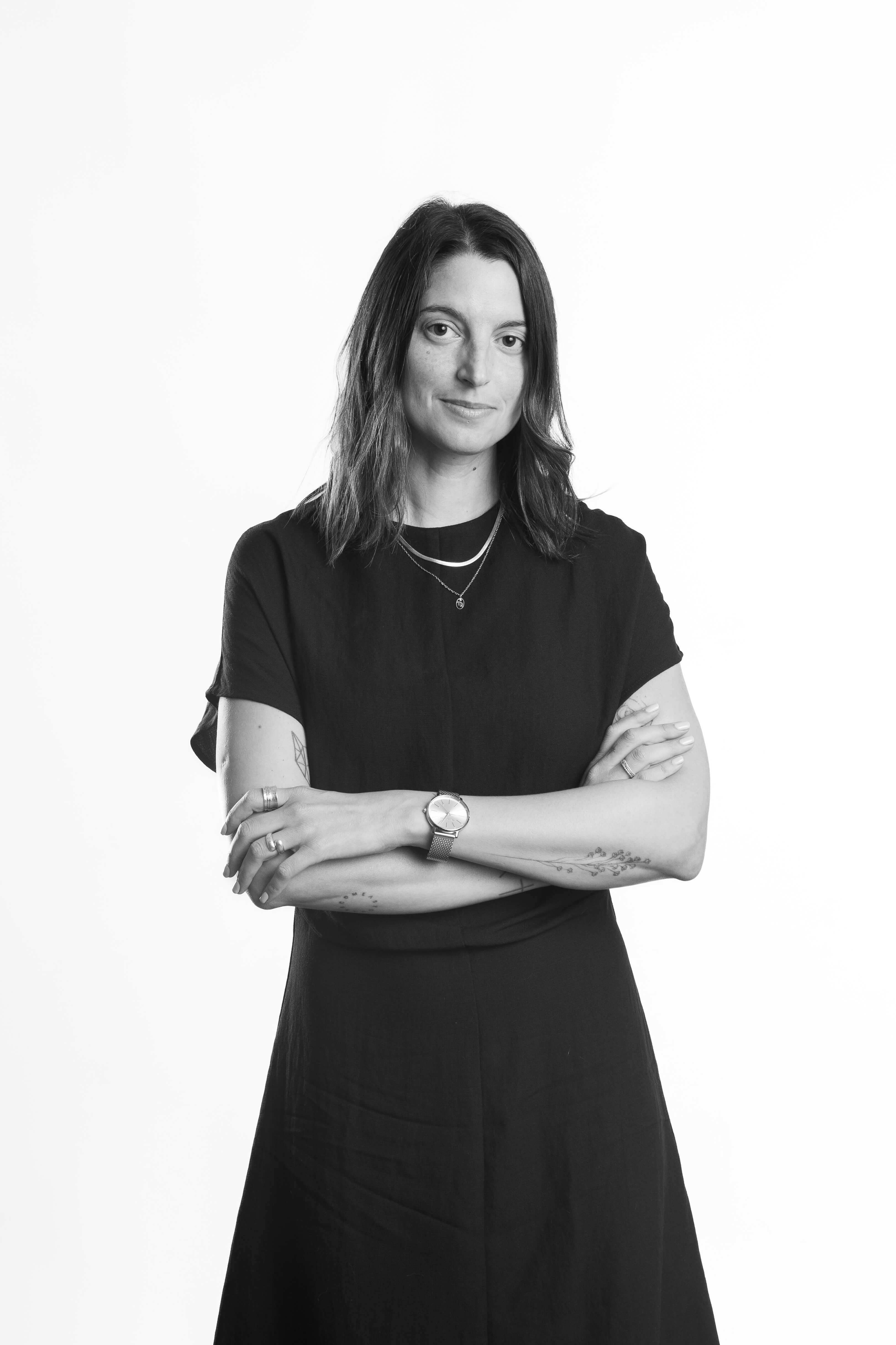 Black and white photo of a woman in a black dress, standing confidently with folded arms. She has shoulder-length hair, tattoos, and wears a watch.