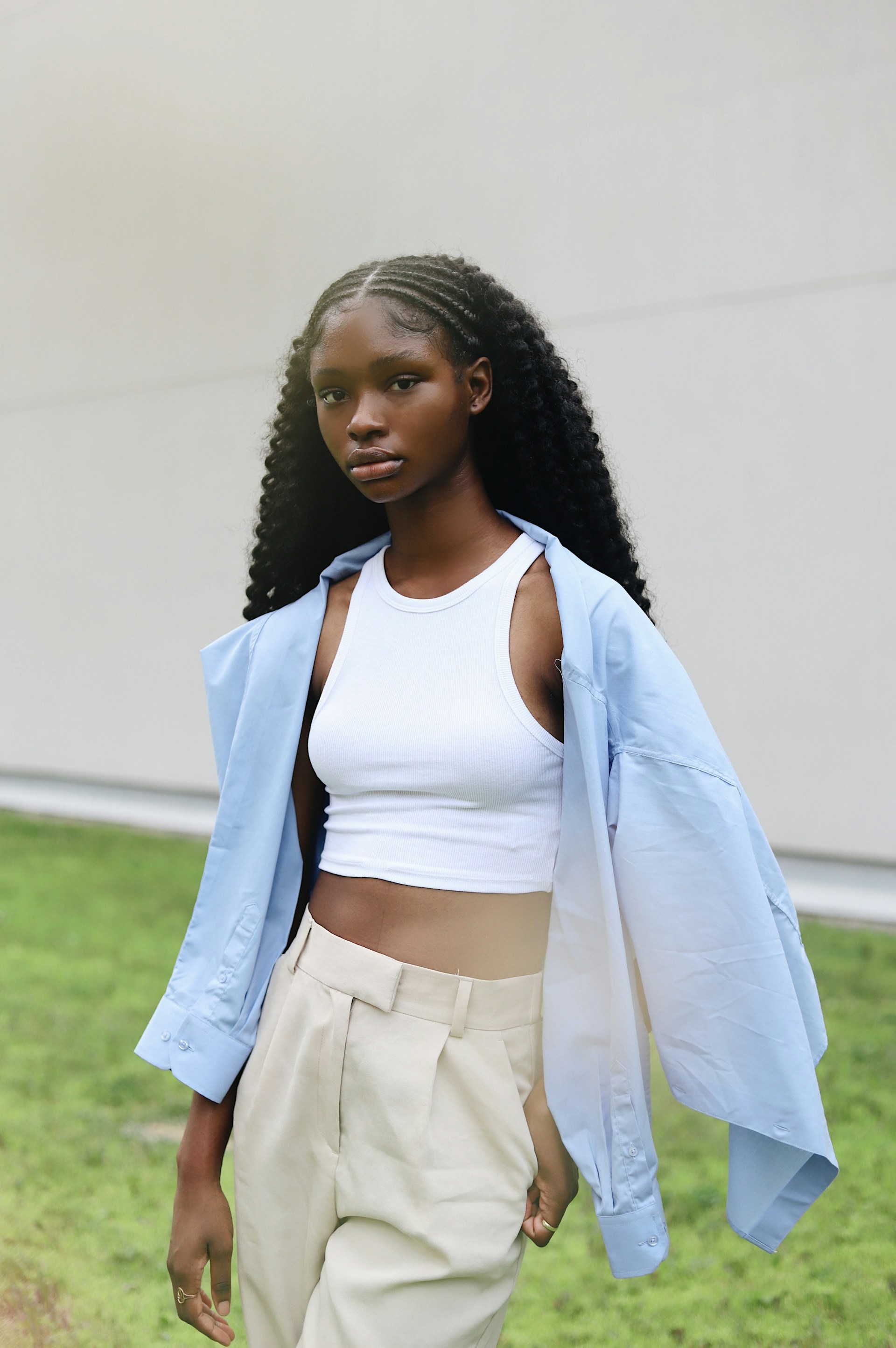 A Black woman standing outdoors against a plain wall and green grass. She is wearing a white crop top, beige pleated trousers, and a light blue shirt draped over her shoulders, looking directly at the camera.