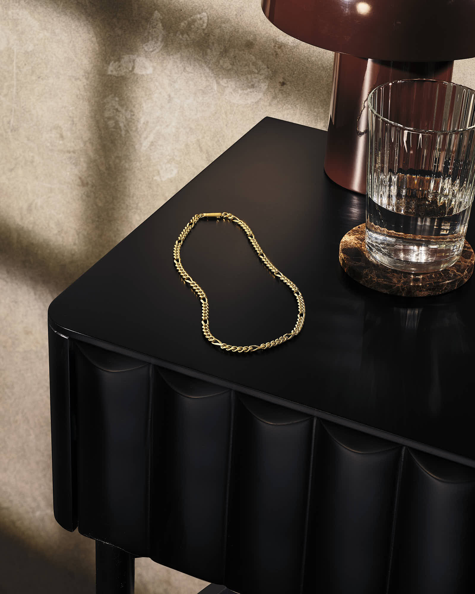 Gold chain necklace on modern black table with glass of water and brown lamp.