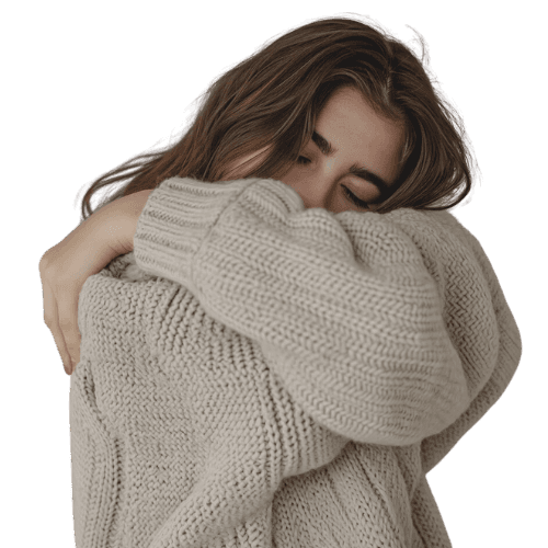 A person hugs themselves while wearing a cozy, light-colored sweater, set against a solid background.