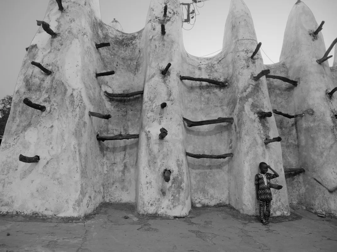 A child standing against the base of a large mud-brick mosque with protruding wooden beams across its curved facade, black and white