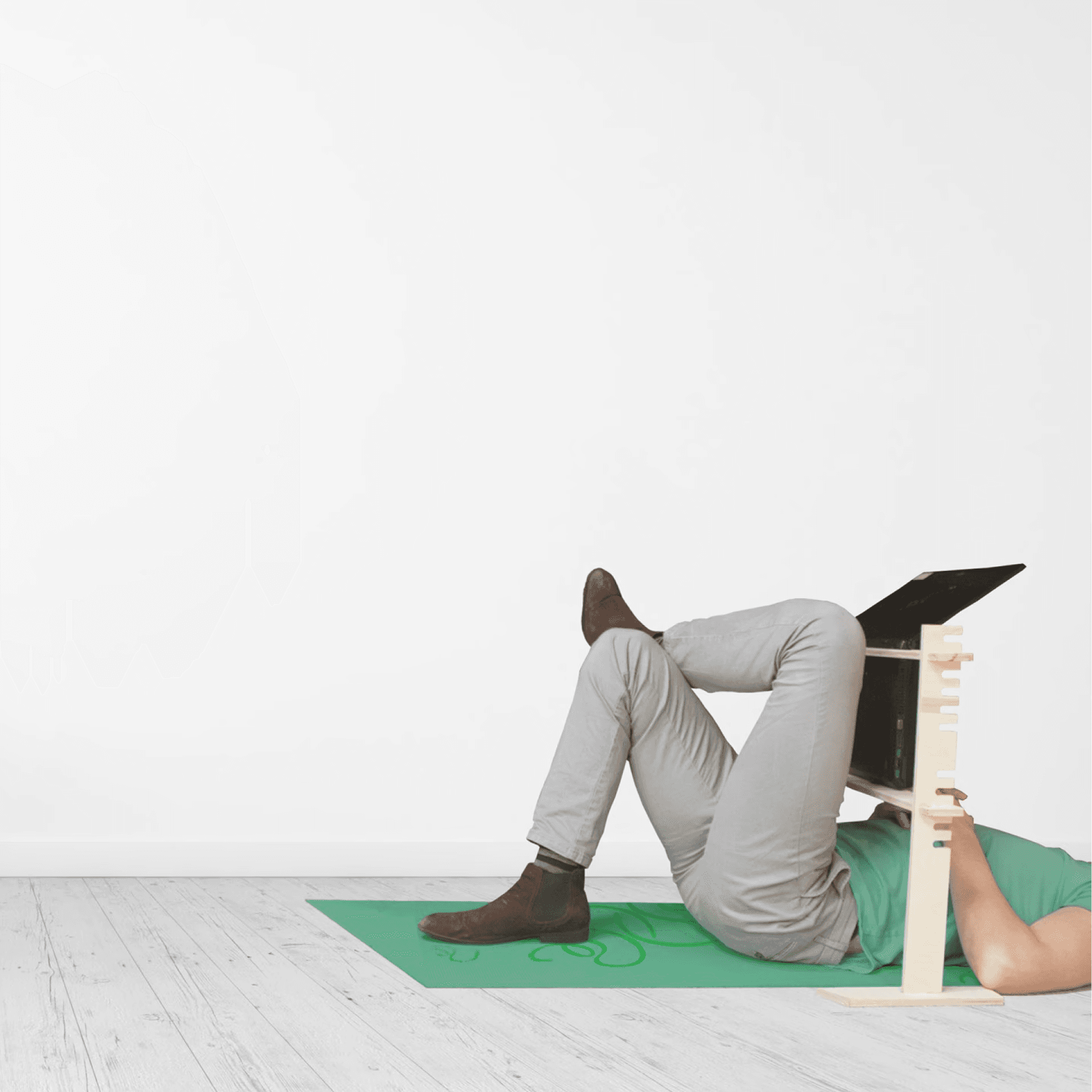 A person using a lie down laptop stand. They are wearing a green tshirt and lying on a green yoga mat. The person, who's head is out of shot, is wearing grey pants. The wall and floor are also grey.