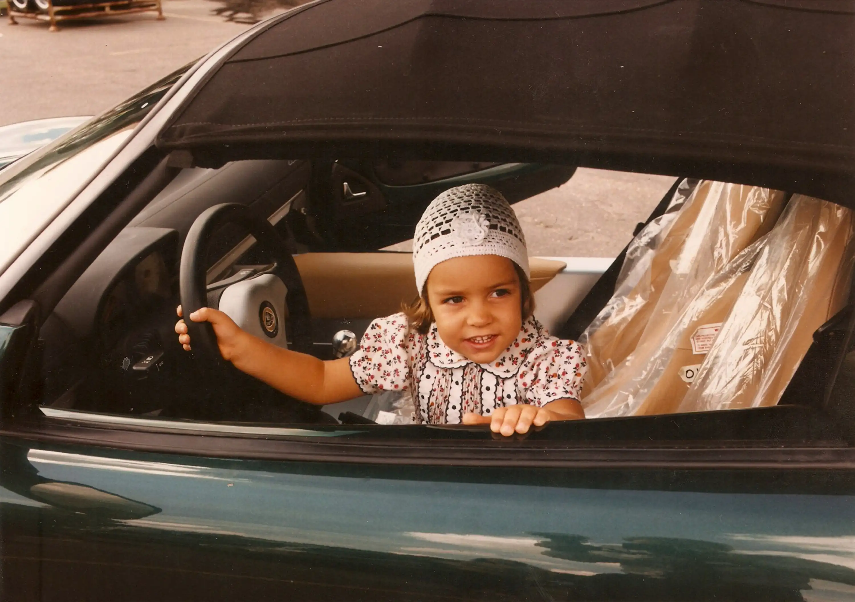 A young Elisa Artioli wearing a floral dress and a crocheted white headscarf sits in the driver’s seat of a green convertible Lotus sports car, smiling and holding the steering wheel.