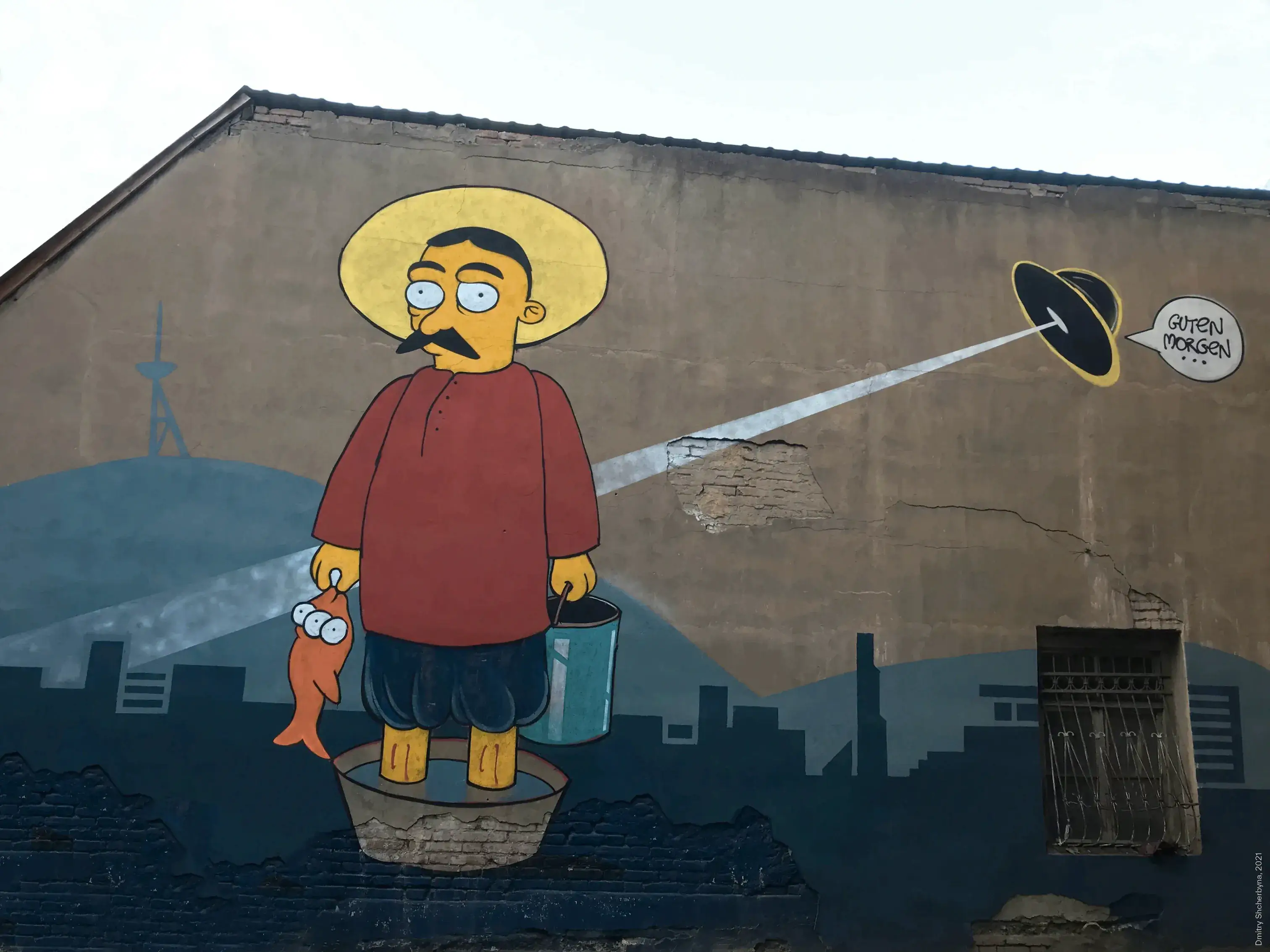 A humorous cartoon mural featuring a mustached man in a yellow hat and red shirt standing in a basin holding an orange fish with a flying saucer beaming light on him, with a speech bubble saying "Guten Morgen."