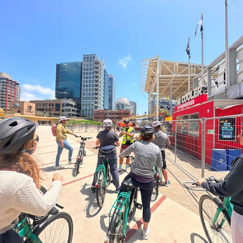 Egy csoport kerékpáros hallgat egy idegenvezetőt egy városi téren magas épületekkel és egy piros kioszkkal derült ég alatt.