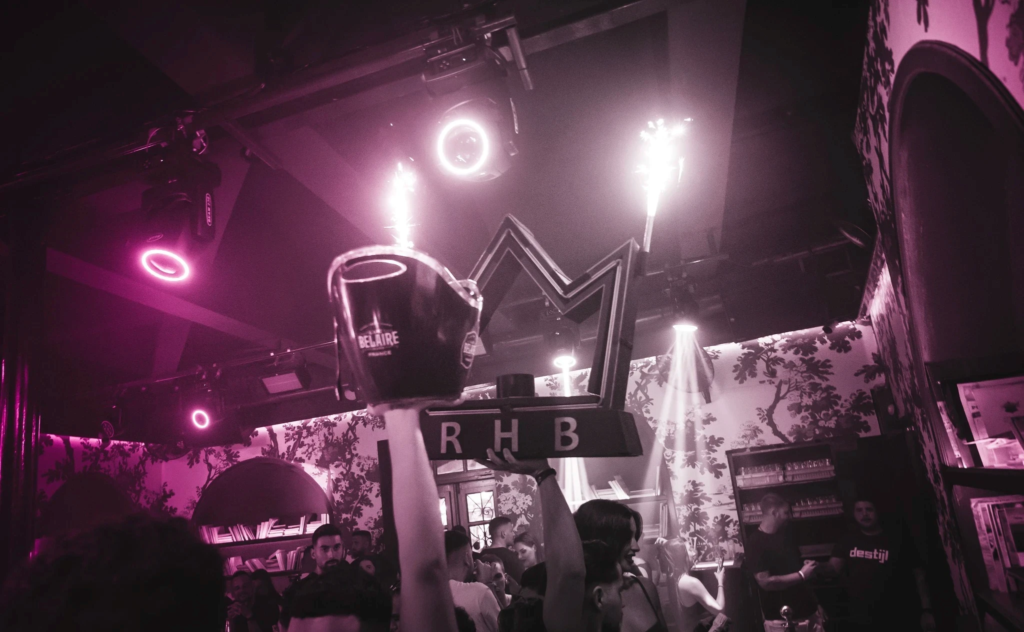A low-angle view from a crowd at the RHB party, showing someone holding up a large "RHB" logo shaped like a crown, alongside a "Belaire" ice bucket, all under bright pink club lights.