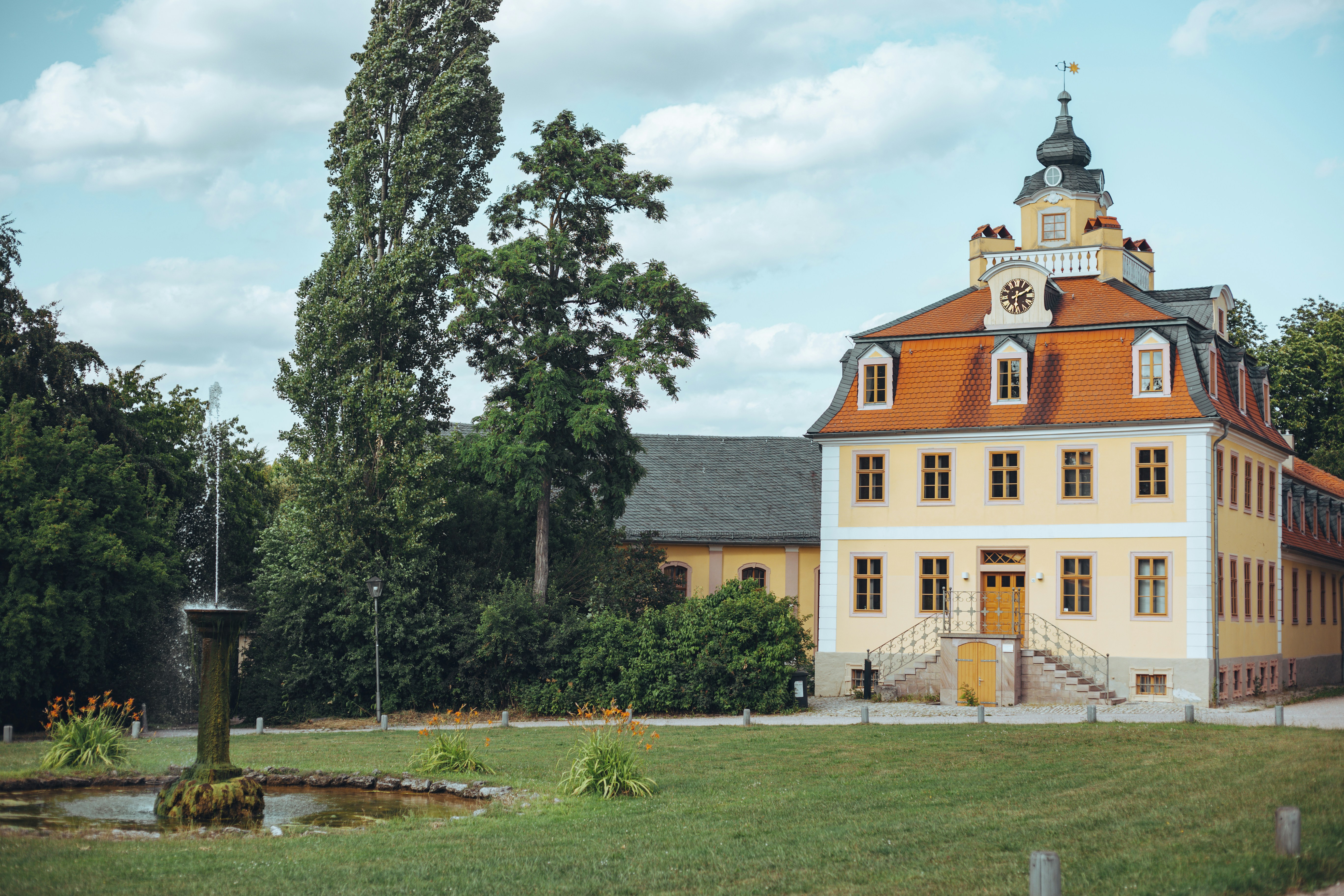 Schloss Belvedere in Weimar, ein barockes Lustschloss mit orangefarbenem Dach und Uhrturm, davor ein Garten mit Springbrunnen.