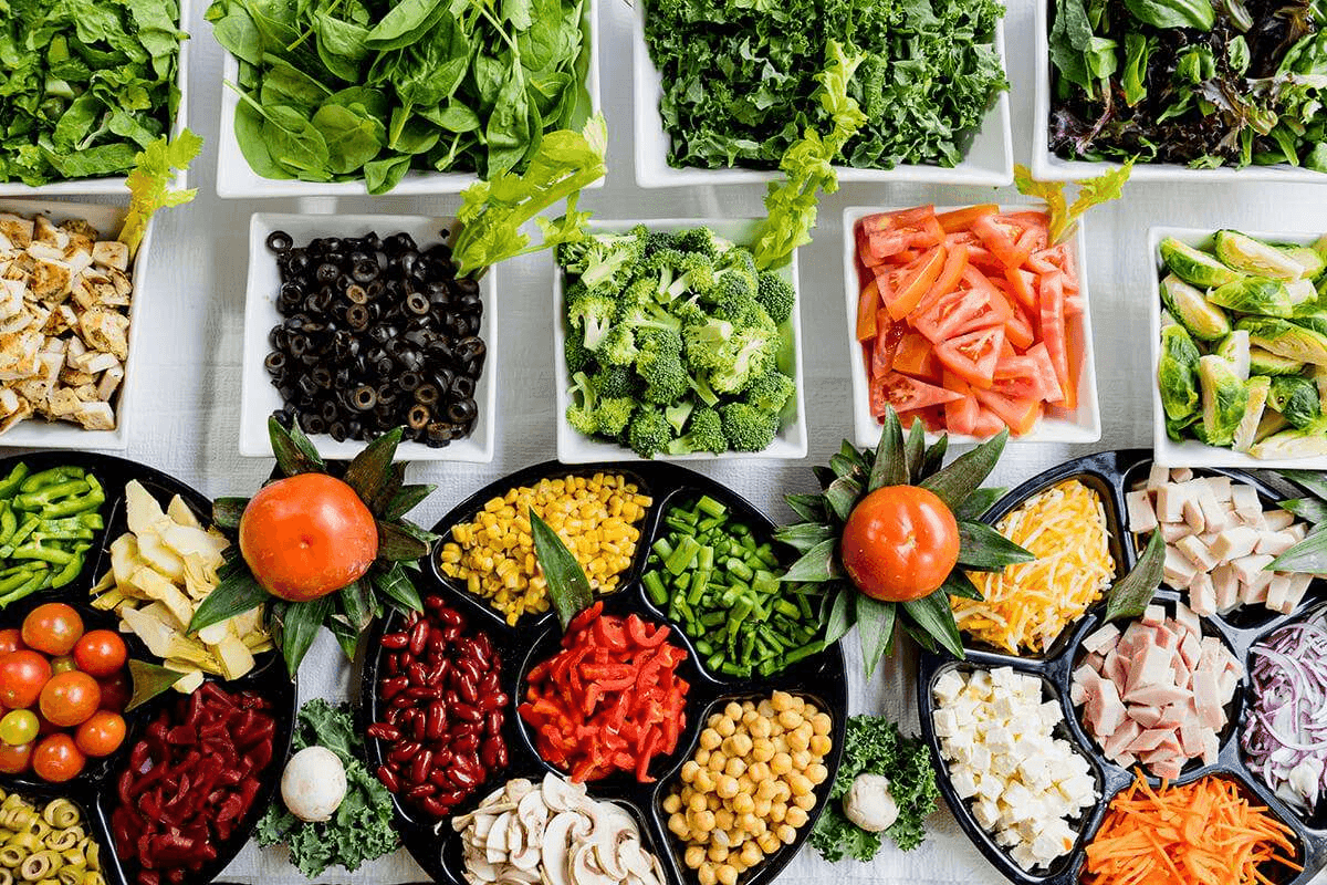 Top-down view of a colorful salad bar featuring various fresh vegetables, greens, beans, and proteins in white and black containers.