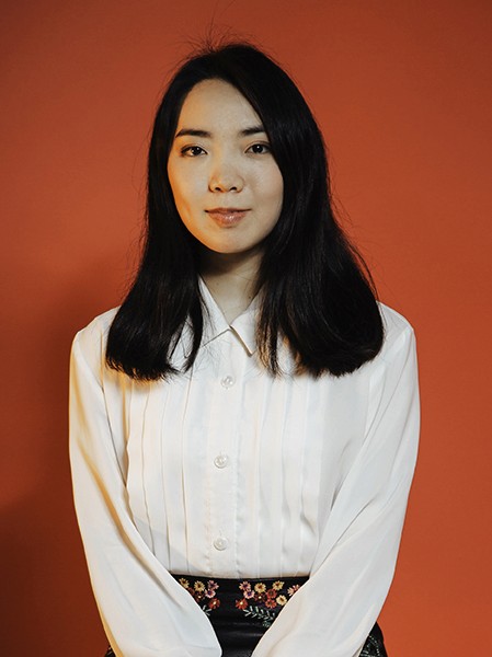 A young woman with long, dark hair stands against a solid orange-brown backdrop. She wears a white long-sleeved button-up shirt with vertical pleats and a collar, buttoned to the top. A black skirt with colorful floral embroidery is visible at the waist. Her expression is neutral, and she looks directly at the camera.