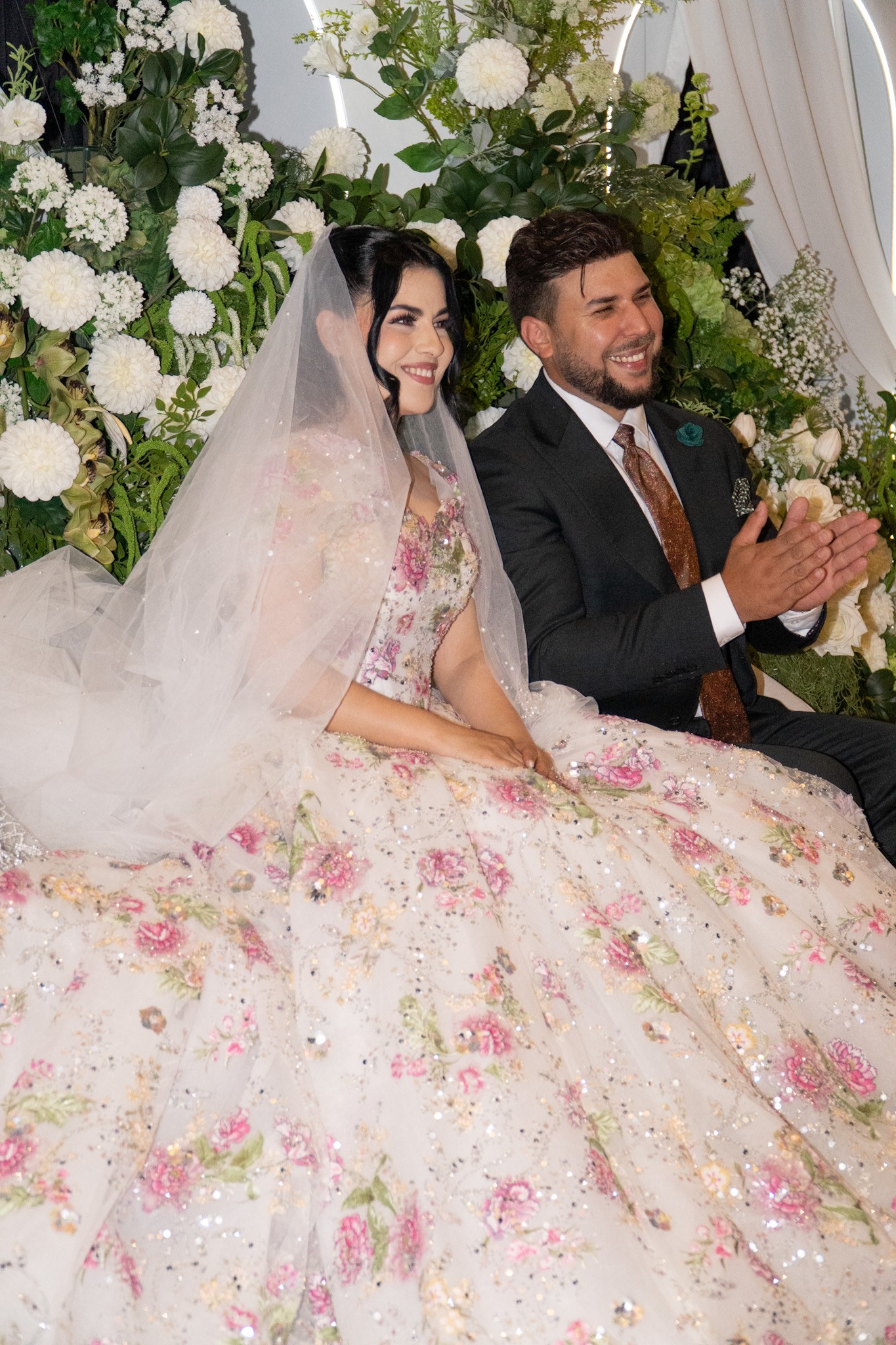 Bride in a floral gown and groom in a dark suit seated together in front of white wedding flowers.