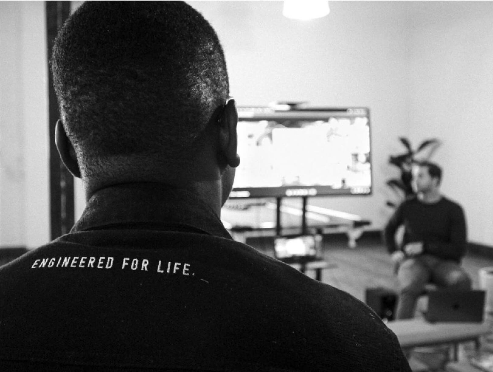 Back view of someone in an "ENGINEERED FOR LIFE." shirt watching a screen with another individual in the room.