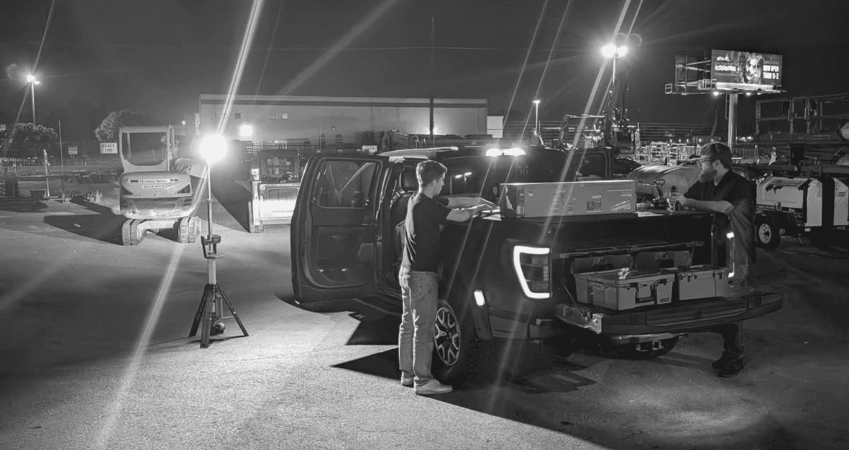 Photo of two men working in an industrial lot with an electronic truck and large floodlights