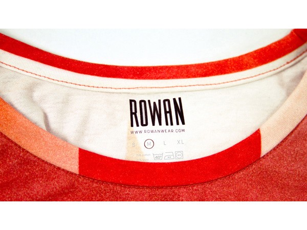 A close-up view of the inside neckline of a red and white Rowan t-shirt. The brand name "ROWAN" is printed in black on the off-white fabric, above a size tag indicating "L" for medium. The neckline features two red bands: a thicker red band at the top and a thinner red band below. The fabric appears soft and slightly textured. The image focuses on the neckline and branding, providing a clear view of the brand and size information.