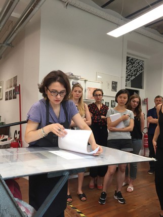 A photo of a group of people looking at a woman how she prepares the paper imprints on a table.