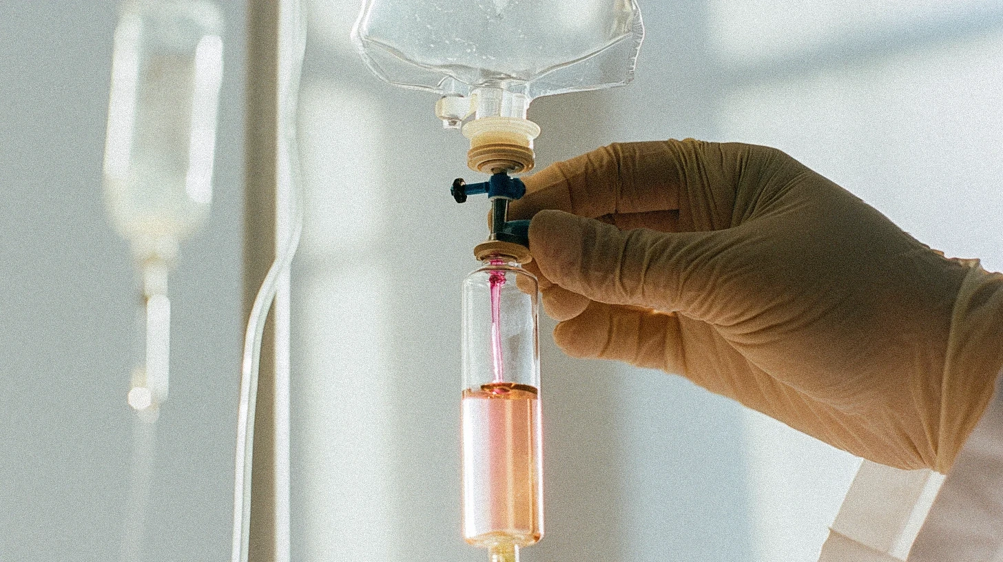 A gloved clinician adjusting the flow valve on an IV drip bag connected to a tinted infusion vial.