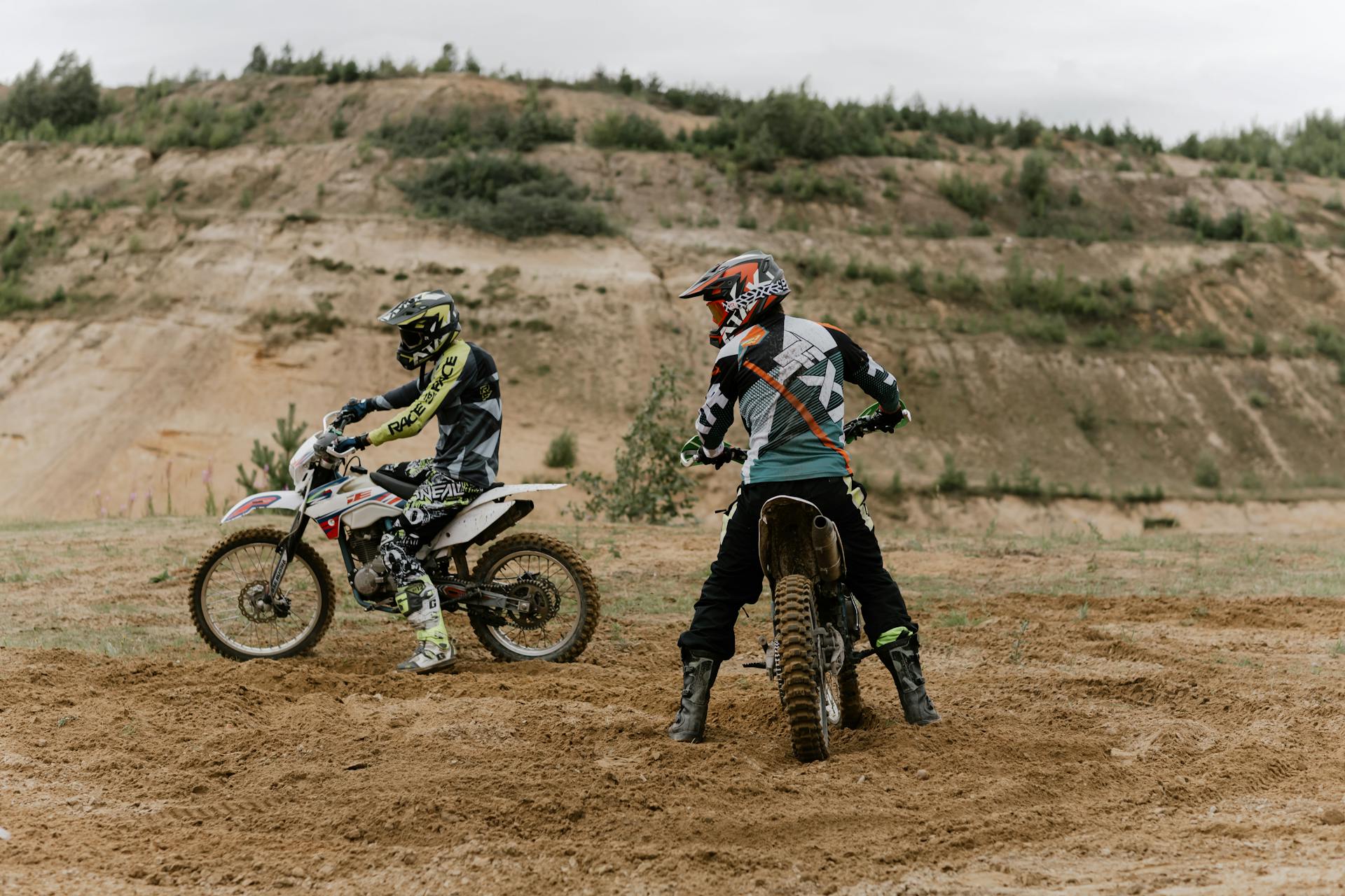 Two motocross riders in full gear and helmets sitting on their dirt bikes in a sandy, open quarry area with hills in the background.