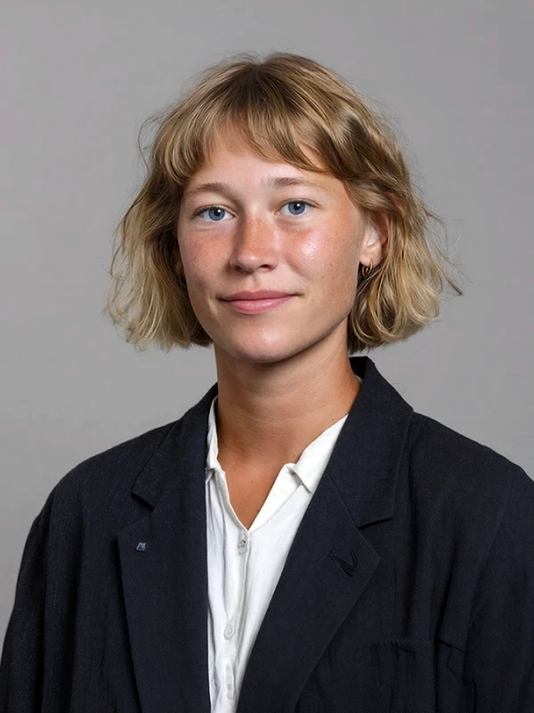 A person with short, wavy hair and a black jacket smiles against a neutral gray background.