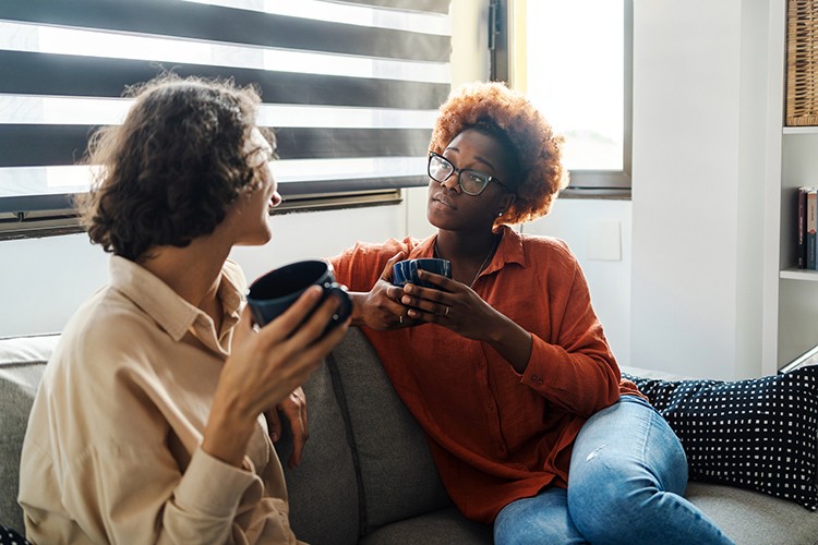 Two women sit on a couch engaged in conversation, each holding a coffee mug. One woman wears a beige shirt while the other, wearing glasses and an orange sweater, sits attentively listening. Natural light streams through striped window blinds behind them, creating an intimate, comfortable setting for what appears to be a meaningful discussion or counseling session.eam of five colleagues smile while gathered around a glass wall covered with colorful sticky notes in pink, yellow, and green. The group appears engaged in collaborative brainstorming or planning, with one woman in the foreground actively placing or organizing notes. The casual, energetic atmosphere suggests creative problem-solving and inclusive teamwork.