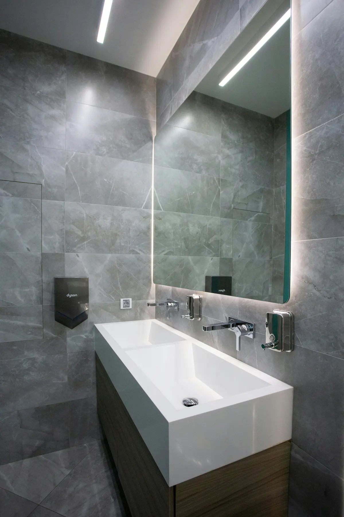 Deeply sanitized modern office restroom featuring a large trough sink, streak-free mirrors with LED backlighting, and polished grey tile walls.
