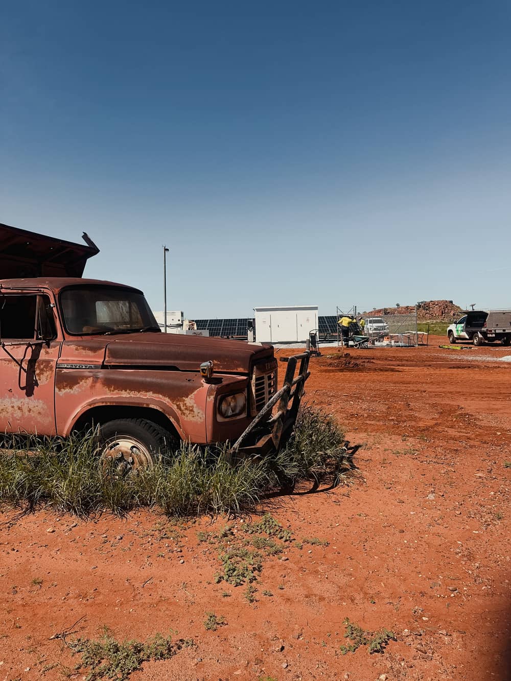 Rusty abandoned truck in foreground with solar panel installation and workers in background on red dirt terrain in Australia