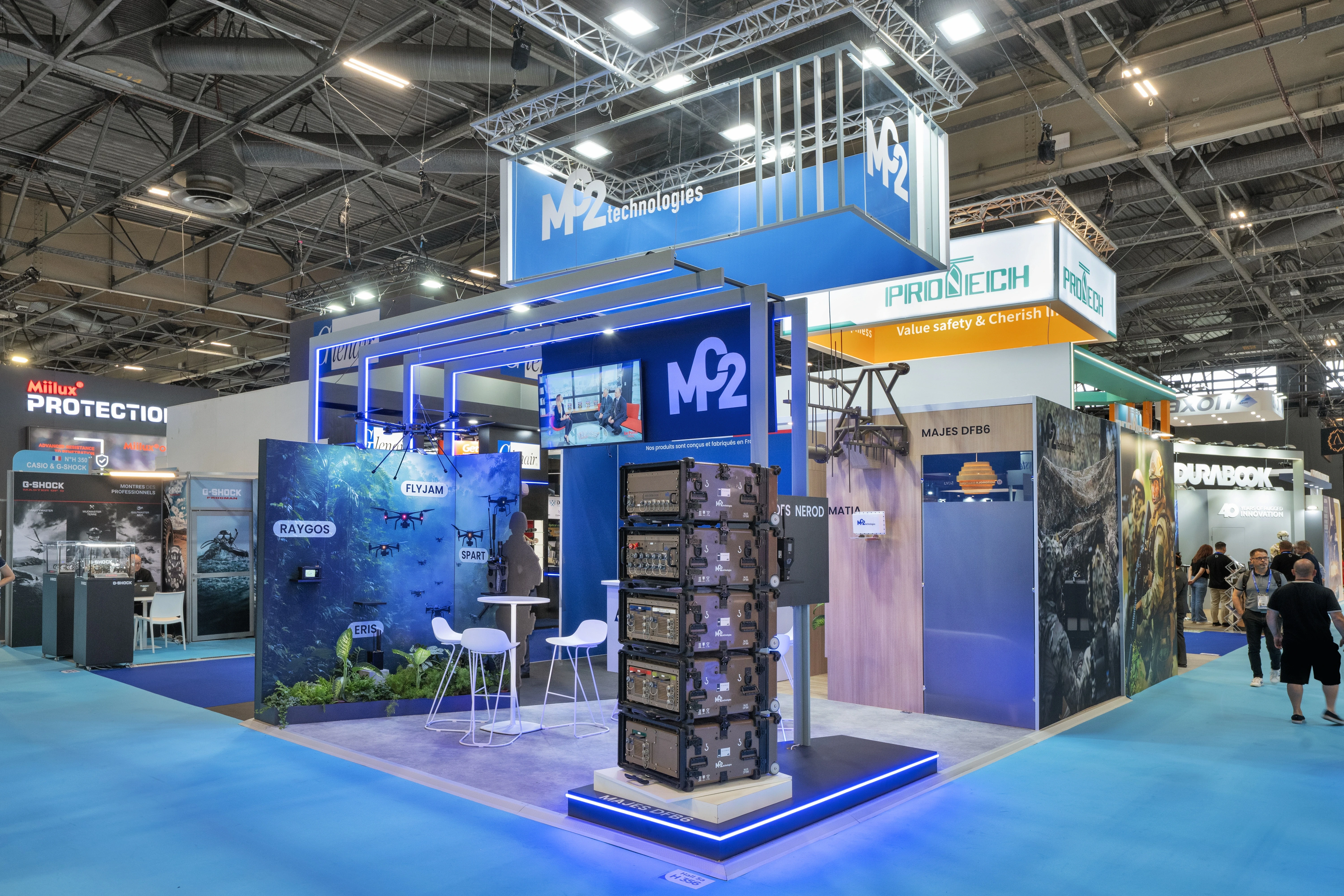 Stand MC2 Technologies lutte anti-drone conçu par EXPACE à Eurosatory 2026, Hall 5A — tour de brouilleurs MAJES DFB6 sur socle lumineux, mur jungle immersif et drones suspendus