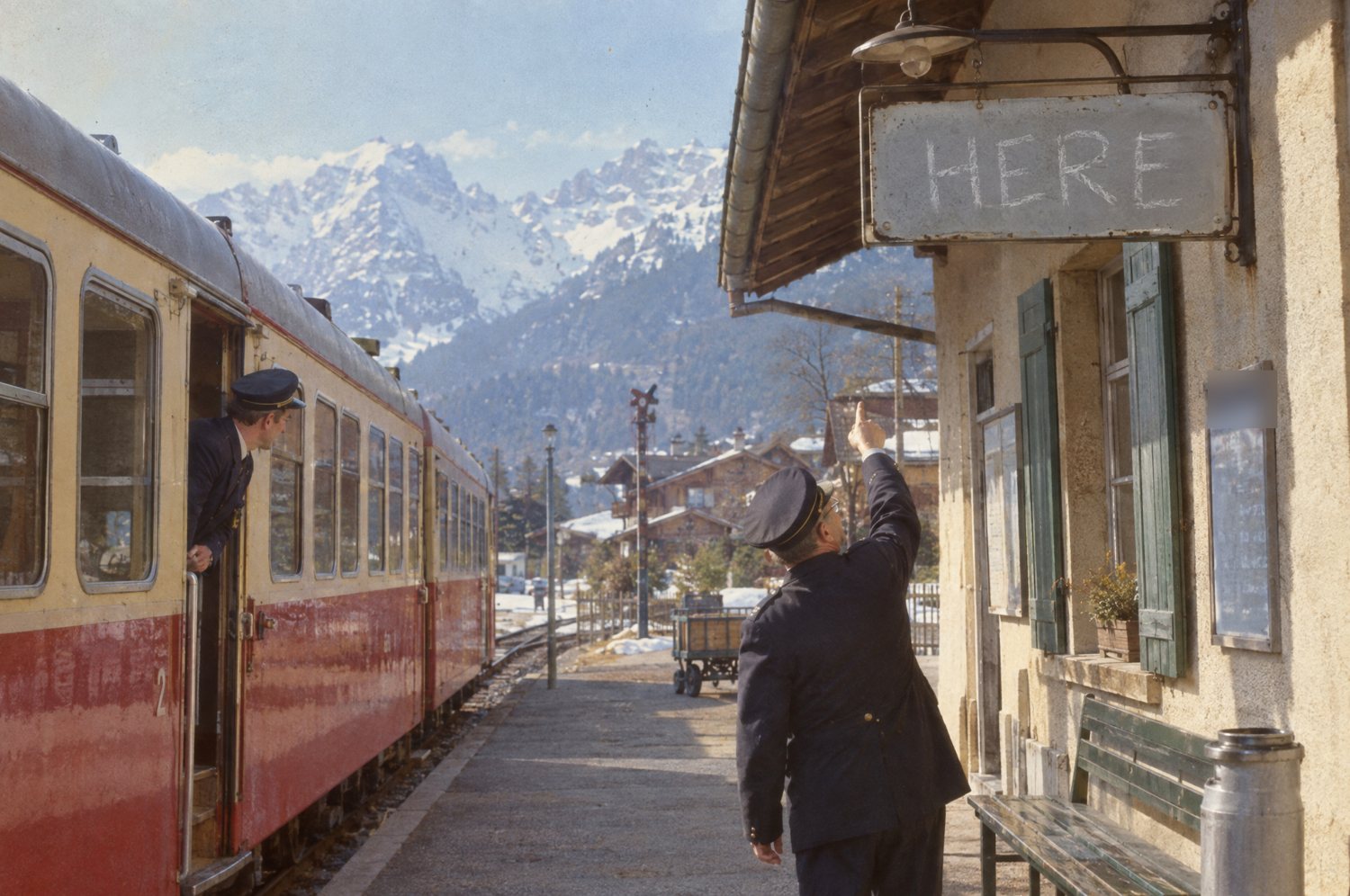 The stationmaster points up at the station sign, now reading HERE in a child's chalk capitals