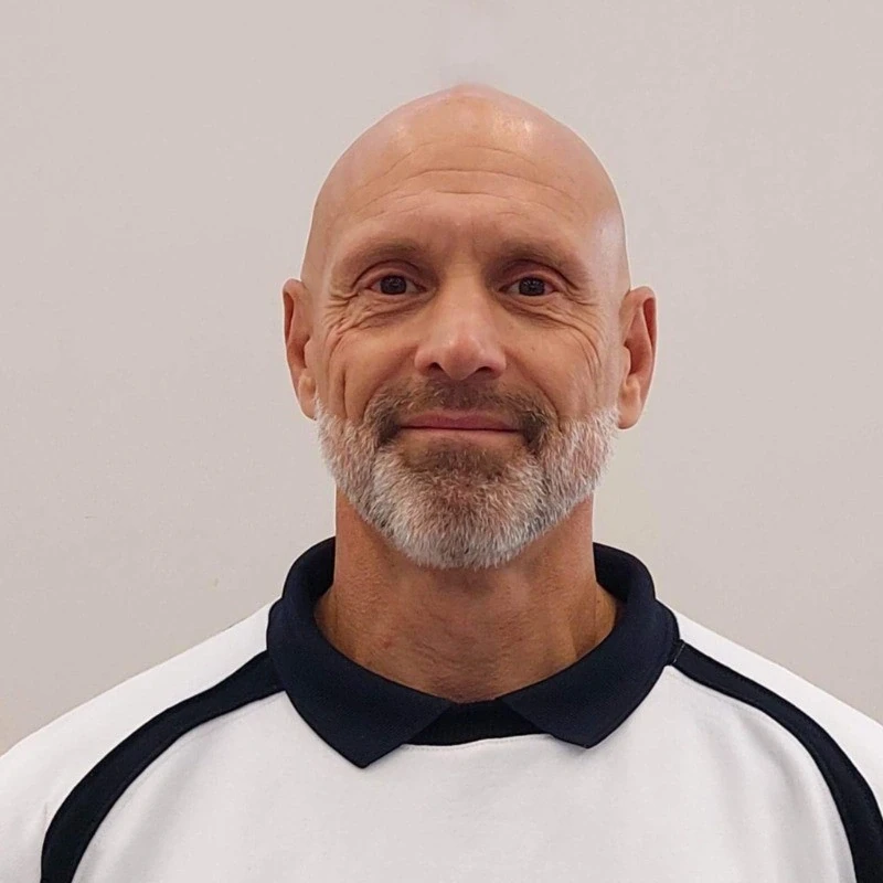 Homme souriant avec barbe blanche portant un polo blanc sur fond neutre.