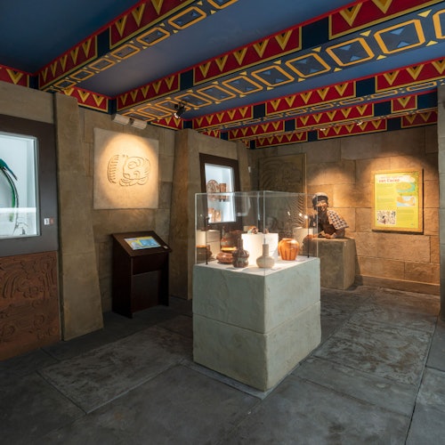 Múzeumi kiállítóterem, régi műtárgyakkal a vitrinben, részletes falfaragványokkal és egy életnagyságú, történelmi öltözetet viselő bábuval.