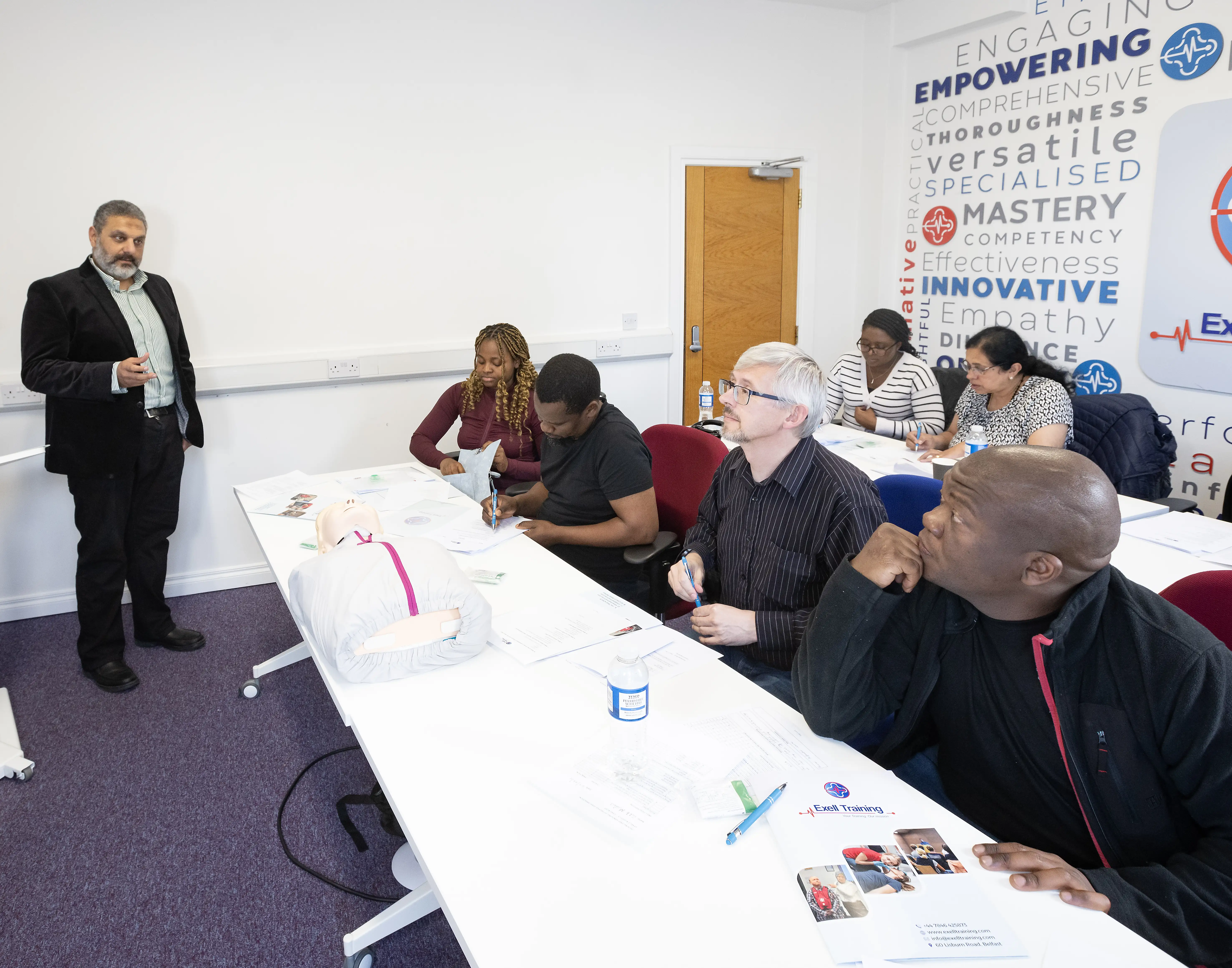 A group of adult learners seated at desks attentively listening to a male instructor during a classroom-based healthcare training session at Exell Training.