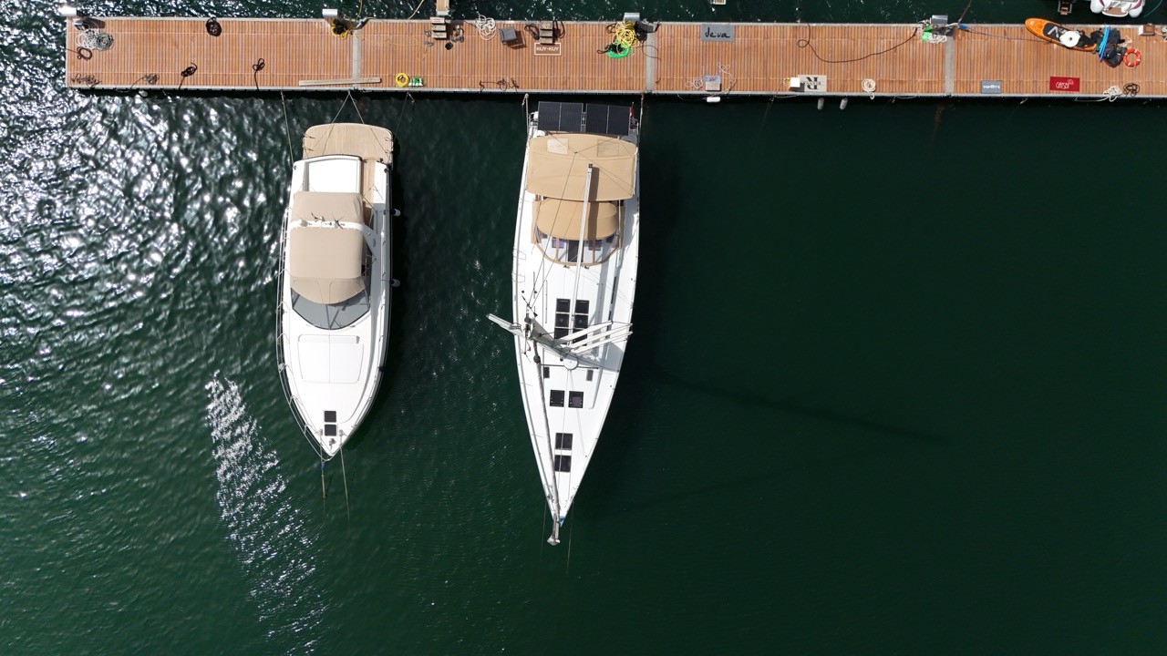 Aerial view looking down on a Hanse 575 sailing yacht with a tan sail cover moored stern-to at a wooden dock, alongside a motor yacht with a tan bimini, in dark green marina water.
