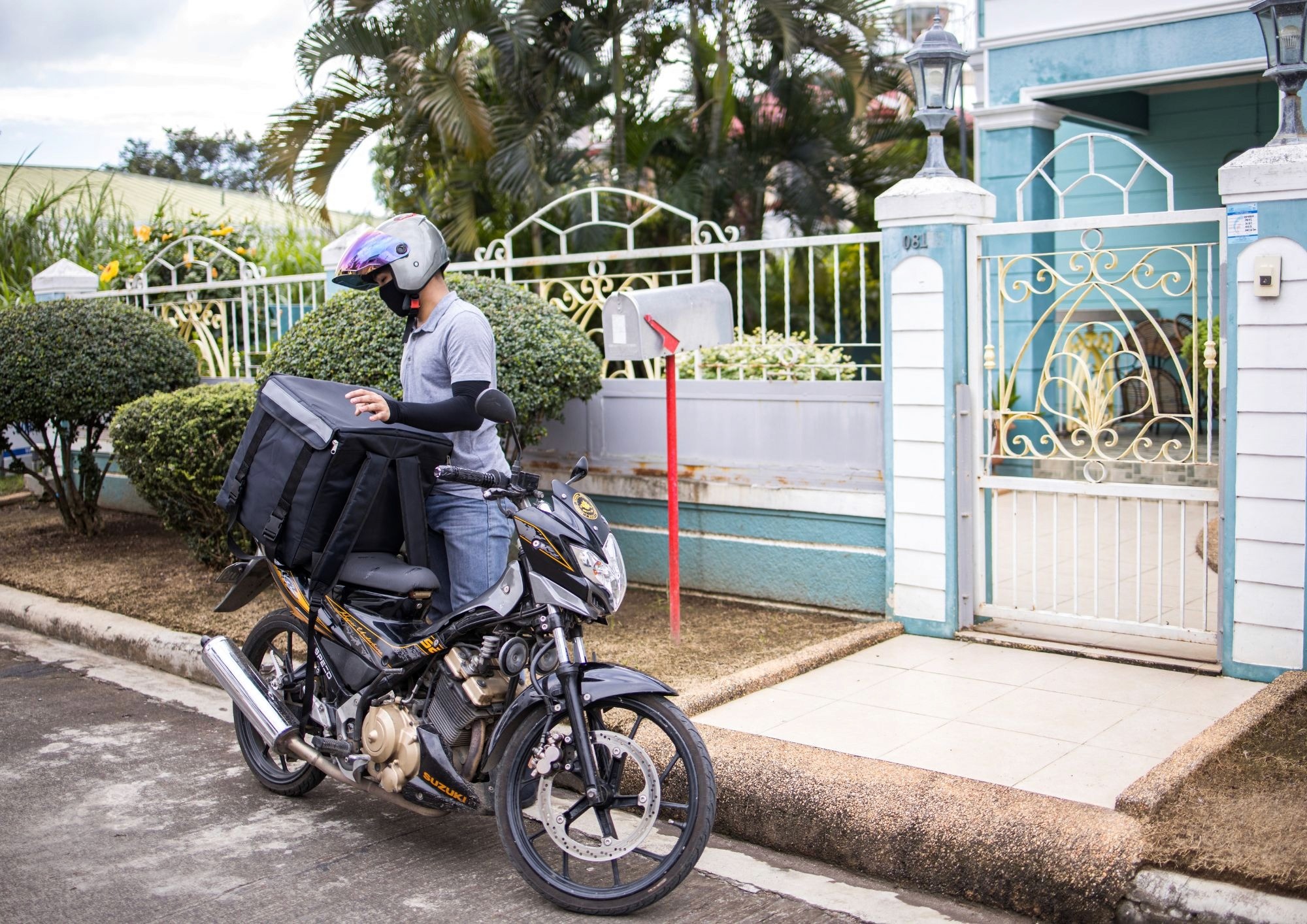 Entregador de moto parado em frente a uma residência segura uma bolsa de delivery ao lado da motocicleta.